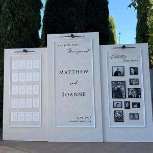 May include: Three cream-colored wedding signs with white frames. The center sign displays the names "MATTHEW and JOANNE" and the date "10.04.2025". The left sign has seating assignments, and the right sign has framed photos.