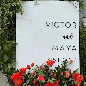 May include: A white sign with the names "VICTOR and MAYA" and the date "05.11.2024" in black lettering. The sign is decorated with vibrant orange flowers and greenery, creating a welcoming display.