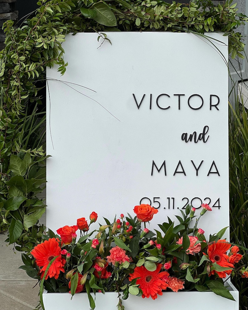 May include: A white sign with the names "VICTOR and MAYA" and the date "05.11.2024" in black lettering. The sign is decorated with vibrant orange flowers and greenery, creating a welcoming display.
