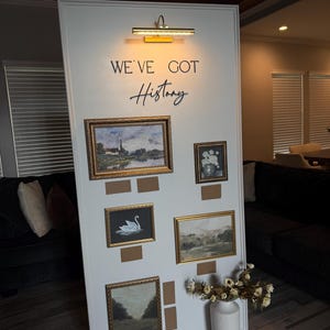 May include: A white display board with framed landscape paintings and the text "WE'VE GOT History". The paintings have gold frames and are mounted on a white board. A white vase with dried flowers is on the floor.