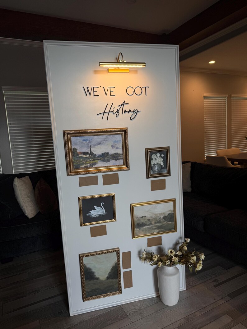 May include: A white display board with framed landscape paintings and the text "WE'VE GOT History". The paintings have gold frames and are mounted on a white board. A white vase with dried flowers is on the floor.