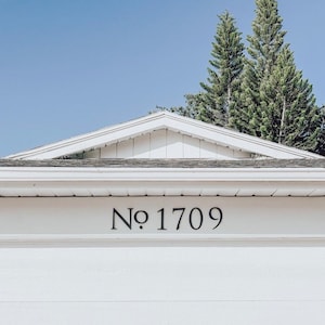 K&ouml;nnte beinhalten: Ein wei&szlig;es Haus mit einem grauen Dach und einer schwarzen Nummerntafel mit der Aufschrift "No 1709". Das Haus steht vor einem strahlend blauen Himmel mit zwei hohen gr&uuml;nen Kiefern im Hintergrund.