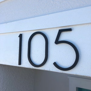 May include: Black metal house number plaque with the number 105 on a white wall.