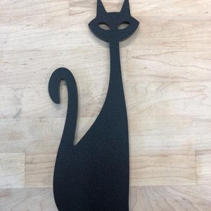 Puede incluir: Silueta negra de un gato con un cuerpo largo y delgado y una cola enroscada. El gato está de pie sobre una superficie de madera clara.