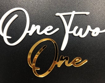 May include: White acrylic script letters spelling "One Two One" with a gold glitter finish on the word "One".