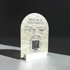 Op de afbeelding: Een boogvormig, parelmoerkleurig bord op een witte voet. Het bevat zwarte tekst met MUST BE 21+ WITH VALID I.D. en Feeling Tipsy? boven een lijntekening van een weckpot met een QR-code en het Venmo-logo onder de pot gedrukt.