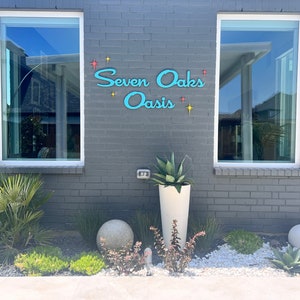May include: A gray brick building with two windows and a blue sign that reads "Seven Oaks Oasis" with yellow stars. There are two round gray planters and a white planter with a green plant in front of the building.