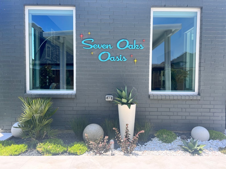 May include: A gray brick building with two windows and a blue sign that reads "Seven Oaks Oasis" with yellow stars. There are two round gray planters and a white planter with a green plant in front of the building.