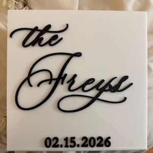 K&ouml;nnte beinhalten: Ein quadratisches wei&szlig;es Acrylschild mit erhabener schwarzer Beschriftung. Der Text lautet "the Freys" in einer Schreibschrift, mit dem Datum "15.02.2026" in einer fetten, serifenlosen Schrift unten mittig auf der glatten Oberfl&auml;che.
