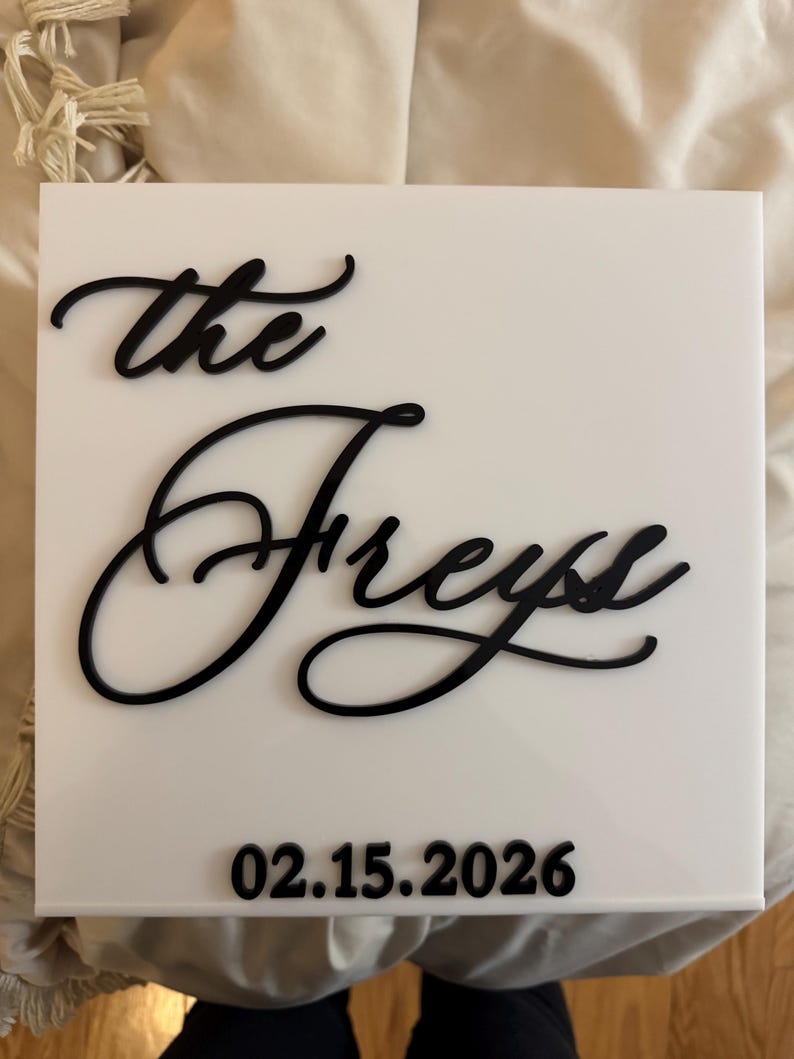 K&ouml;nnte beinhalten: Ein quadratisches wei&szlig;es Acrylschild mit erhabener schwarzer Beschriftung. Der Text lautet "the Freys" in einer Schreibschrift, mit dem Datum "15.02.2026" in einer fetten, serifenlosen Schrift unten mittig auf der glatten Oberfl&auml;che.