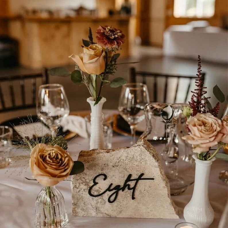 May include: A rustic wedding table setting with a stone table number that reads "Eight". The table is decorated with flowers in shades of pink, peach, and burgundy, and clear glass vases.
