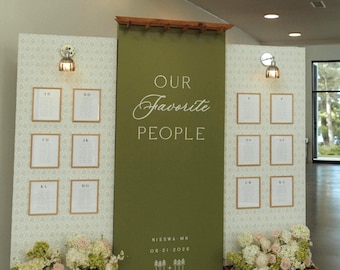 Puede incluir: Un expositor de mesa de boda de tres paneles con un tablero central verde oliva con el texto OUR Favorite PEOPLE, NISSWA MN, 06-21-2026 e iconos de árboles. Los paneles laterales tienen papel pintado blanco, lámparas y listas de invitados.