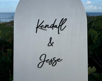 Könnte beinhalten: Ein weißes, bogenförmiges Holzschild mit den Namen "Kendall & Jesse" in schwarzer Schrift. Das Schild befindet sich im Freien, mit Blick auf das Meer und den Himmel im Hintergrund. Das Schild ist aus Holz.