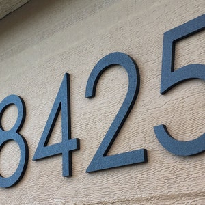 Puede incluir: Números de casa metálicos gris oscuro 18425 montados en una pared beige texturizada. Los números están en una fuente sans-serif moderna, con un acabado ligeramente texturizado, creando un contraste sutil.