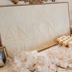 May include: A display featuring a sign with the name "ANTON GIOVANNI" in embossed letters. The sign is above a table with cupcakes and wrapped treats, suggesting a celebration or event. A gold framed mirror is above the sign.