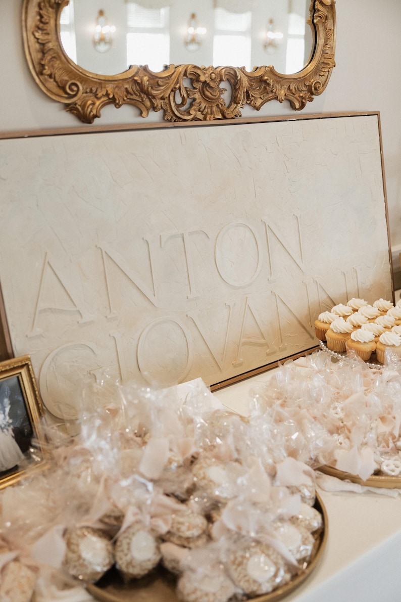 May include: A display featuring a sign with the name "ANTON GIOVANNI" in embossed letters. The sign is above a table with cupcakes and wrapped treats, suggesting a celebration or event. A gold framed mirror is above the sign.