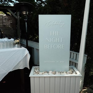 May include: A light blue sign with the words "Celebrating THE NIGHT BEFORE SEPTEMBER 19, 2025" in embossed lettering. The sign is set in a white planter filled with rocks, with a wooden deck and greenery in the background.