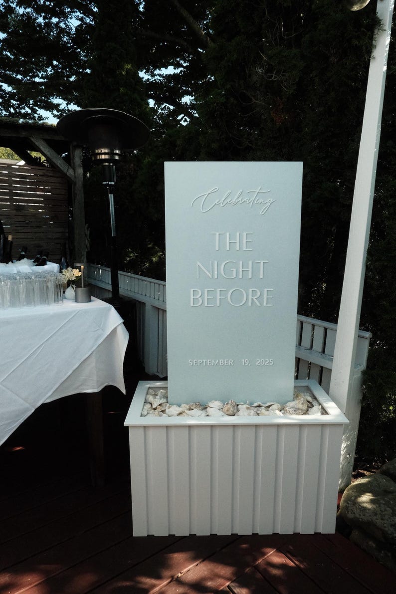 May include: A light blue sign with the words "Celebrating THE NIGHT BEFORE SEPTEMBER 19, 2025" in embossed lettering. The sign is set in a white planter filled with rocks, with a wooden deck and greenery in the background.