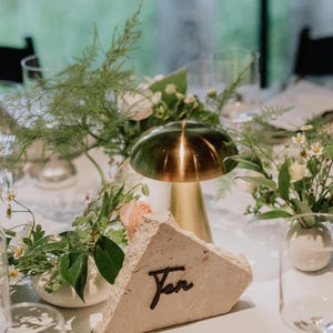 May include: A rustic, triangular table number made of light beige stone with the word "Ten" written in black script. The table number is sitting on a white tablecloth with a gold metal dome and green floral arrangements.