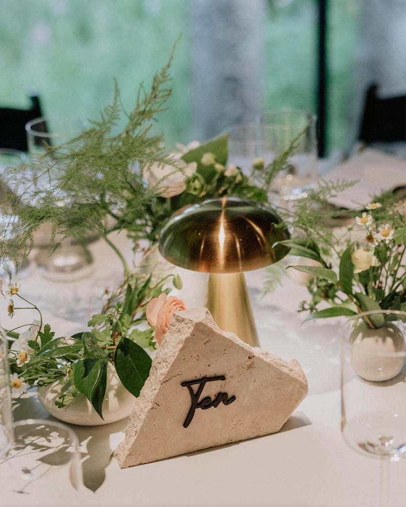 May include: A rustic, triangular table number made of light beige stone with the word "Ten" written in black script. The table number is sitting on a white tablecloth with a gold metal dome and green floral arrangements.