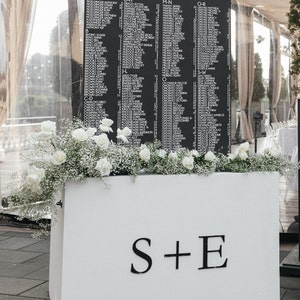 May include: A black sign with white lettering that reads "OUR favorite PEOPLE" lists names in alphabetical order. The sign is decorated with white roses and baby's breath and sits atop a white box with the letters "S+E".