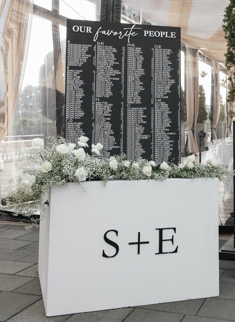 May include: A black sign with white lettering that reads "OUR favorite PEOPLE" lists names in alphabetical order. The sign is decorated with white roses and baby's breath and sits atop a white box with the letters "S+E".
