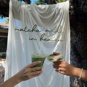 K&ouml;nnte beinhalten: Ein durchsichtiger Plastikbecher mit einem gr&uuml;nen Matcha-Getr&auml;nk und eine gerippte Glasschale mit einem geschichteten Dessert, das mit einer Erdbeere garniert ist, vor einem wei&szlig;en Stoffhintergrund mit dem Text matcha made in heaven.