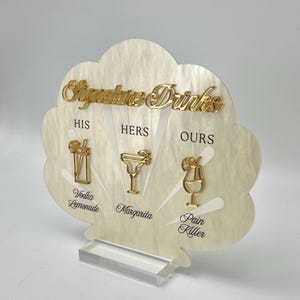 Puede incluir: Un letrero decorativo para bar con las palabras "Signature Drinks" en letras doradas. El letrero tiene forma de concha de vieira y presenta ilustraciones doradas de cócteles para las bebidas "His", "Hers" y "Ours": Vodka Lemonade, Margarita y Pain Killer.