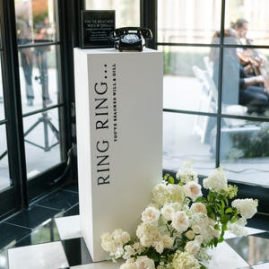 May include: A white pedestal with the text "RING RING... YOU'VE REACHED WILL & GILL" supports a black rotary phone and a sign. White flowers cascade from the base. The scene is set against a backdrop of large windows.