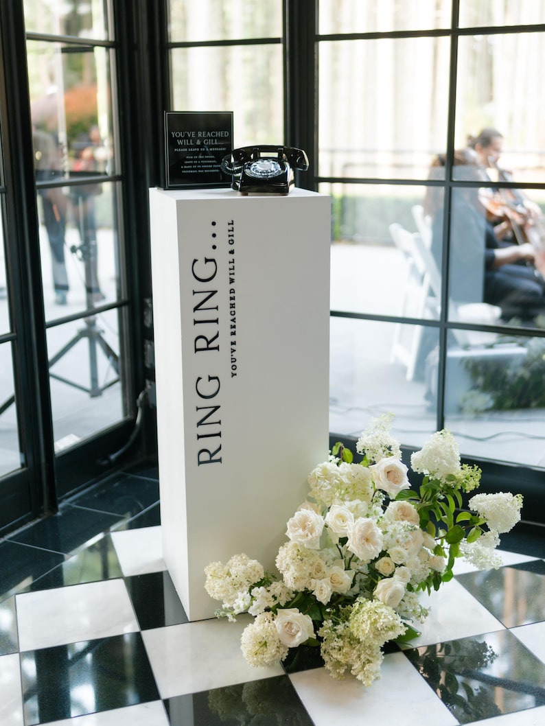May include: A white pedestal with the text "RING RING... YOU'VE REACHED WILL & GILL" supports a black rotary phone and a sign. White flowers cascade from the base. The scene is set against a backdrop of large windows.