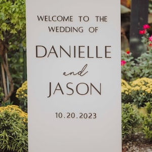 May include: A white rectangular wedding sign with the text "WELCOME TO THE WEDDING OF DANIELLE and JASON 10.20.2023" in dark brown lettering. The sign is surrounded by greenery and flowers.