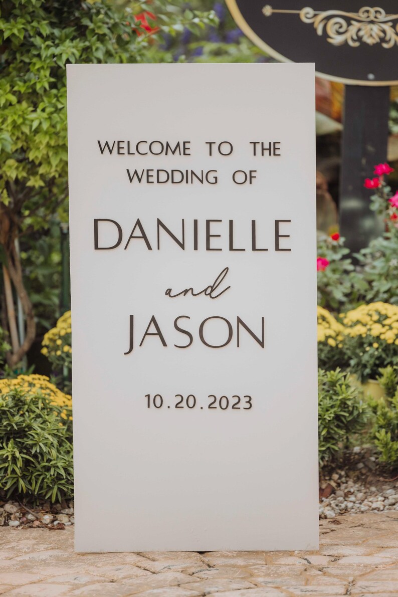 May include: A white rectangular wedding sign with the text "WELCOME TO THE WEDDING OF DANIELLE and JASON 10.20.2023" in dark brown lettering. The sign is surrounded by greenery and flowers.