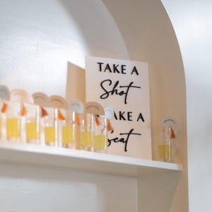 K&ouml;nnte beinhalten: Ein wei&szlig;es rechteckiges Schild mit schwarzer Aufschrift "TAKE A SHOT TAKE A SEAT", platziert neben einer Reihe klarer Schnapsgl&auml;ser mit gelber Fl&uuml;ssigkeit, die jeweils mit einem kleinen runden Papieranh&auml;nger und einer orangefarbenen Quaste versehen sind.