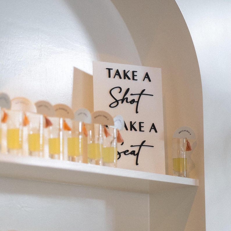 K&ouml;nnte beinhalten: Ein wei&szlig;es rechteckiges Schild mit schwarzer Aufschrift "TAKE A SHOT TAKE A SEAT", platziert neben einer Reihe klarer Schnapsgl&auml;ser mit gelber Fl&uuml;ssigkeit, die jeweils mit einem kleinen runden Papieranh&auml;nger und einer orangefarbenen Quaste versehen sind.