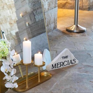 May include: A gold-colored metal candle holder with three white candles, two lit. The holder sits on a matching oval tray. A white triangular sign with the text "THE MERCIAS" is also present.
