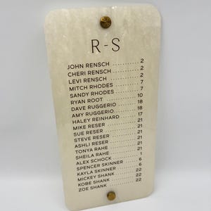 May include: A rectangular, off-white, marbled acrylic seating chart sign with rounded corners. The sign features the text R-S at the top, followed by a list of names and corresponding table numbers. It is secured by two gold-toned metal bolts.