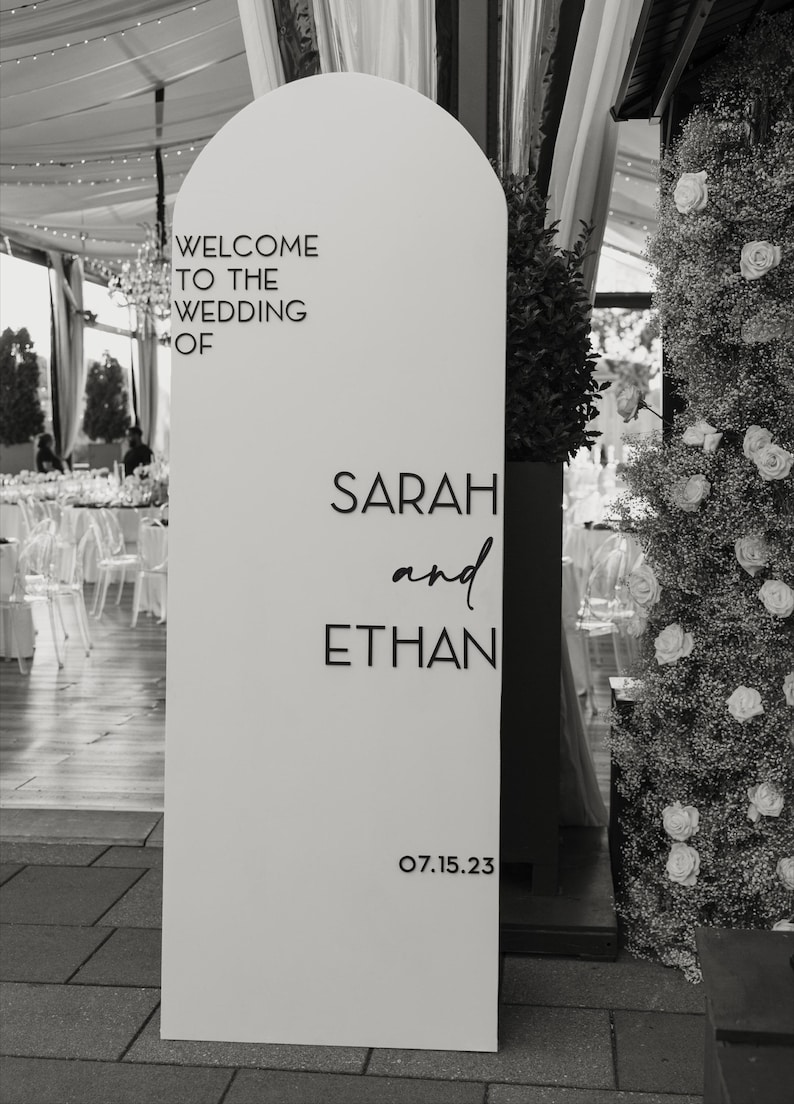 May include: A white wedding welcome sign with an arched top. The sign reads "WELCOME TO THE WEDDING OF SARAH and ETHAN" with the date "07.15.23" at the bottom. The background includes a floral arrangement and a wedding reception.