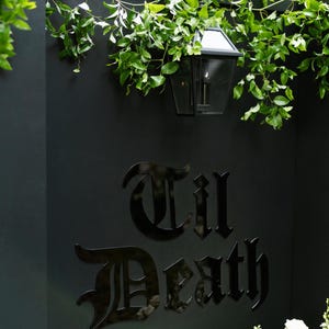 K&ouml;nnte beinhalten: Eine mattschwarze Wand mit dem gl&auml;nzend schwarzen Schriftzug 'Til Death' in gotischer Schrift. &Uuml;ber dem Text h&auml;ngt eine schwarze Metalllaterne, gr&uuml;ne Ranken h&auml;ngen &uuml;ber die Oberkante der Wand und wei&szlig;e Blumen sind unten rechts zu sehen.