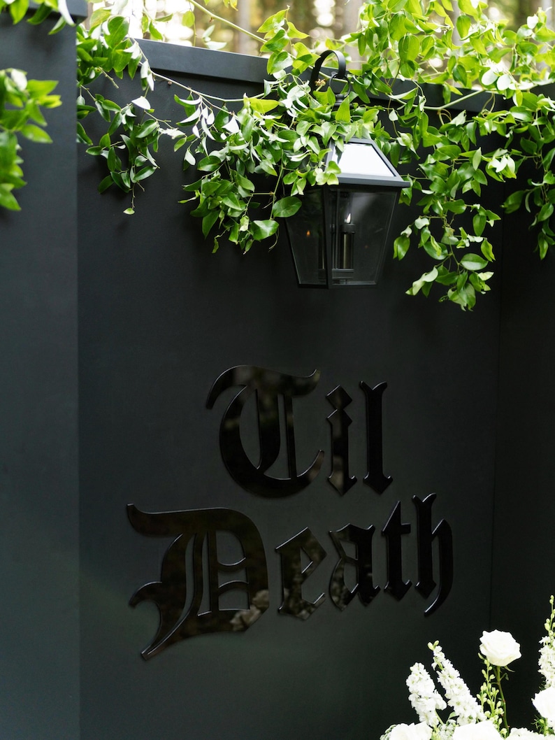 K&ouml;nnte beinhalten: Eine mattschwarze Wand mit dem gl&auml;nzend schwarzen Schriftzug 'Til Death' in gotischer Schrift. &Uuml;ber dem Text h&auml;ngt eine schwarze Metalllaterne, gr&uuml;ne Ranken h&auml;ngen &uuml;ber die Oberkante der Wand und wei&szlig;e Blumen sind unten rechts zu sehen.