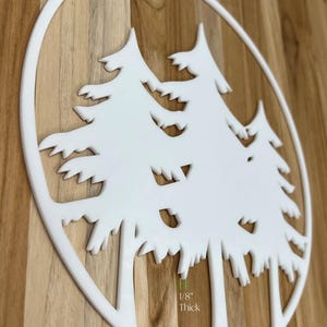 K&ouml;nnte beinhalten: Eine wei&szlig;e, kreisf&ouml;rmige Wanddekoration mit einem Ausschnittdesign aus drei Kiefern. Das St&uuml;ck ist von einem d&uuml;nnen wei&szlig;en Rand umrahmt und 0,32 cm dick. Das Design besteht aus klaren Linien und scharfen Kanten, die eine Waldsilhouette bilden.