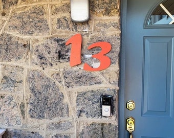 Könnte beinhalten: Eine rote Hausnummer "13" ist an einer Steinwand angebracht. Unterhalb der Nummer ist eine schwarze Türklingelkamera montiert. Rechts von der Nummer befindet sich eine blaue Tür mit einem goldenen Griff.