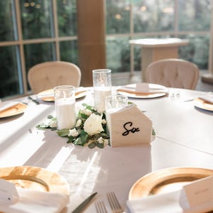 May include: A white table setting with gold chargers, white napkins, and silver cutlery. A white marble table number with the word "Six" written in black is in the center of the table, surrounded by white flowers and candles.