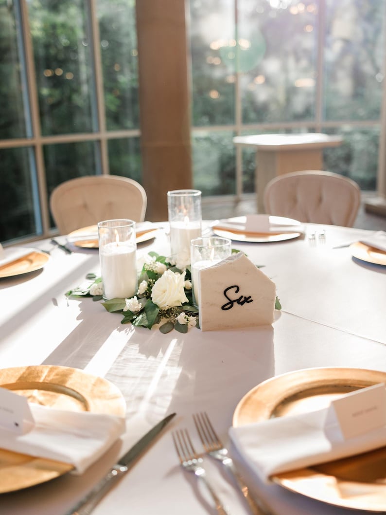 May include: A white table setting with gold chargers, white napkins, and silver cutlery. A white marble table number with the word "Six" written in black is in the center of the table, surrounded by white flowers and candles.