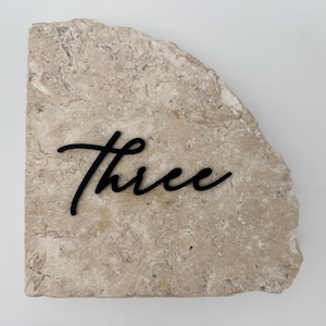 May include: A beige, irregularly shaped stone with the word "Three" in black cursive script. The stone has a rough, textured surface and a natural, organic appearance. The text is centered on the stone.