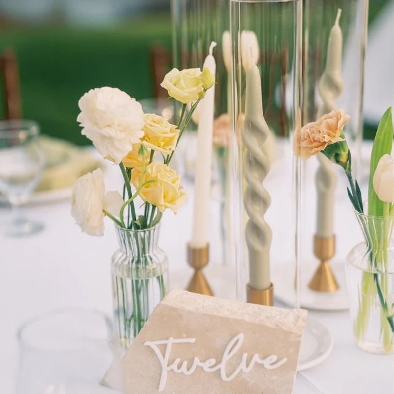 May include: A table setting with a white tablecloth, a beige stone place card with the word "Twelve" written in white, and a small vase with yellow and white flowers. There are also two tall glass vases with candles.