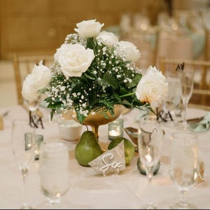 May include: A white floral centerpiece with white roses and baby's breath in a gold bowl. The centerpiece is on a white tablecloth with green placemats and clear glass glasses. The table number is "Six".