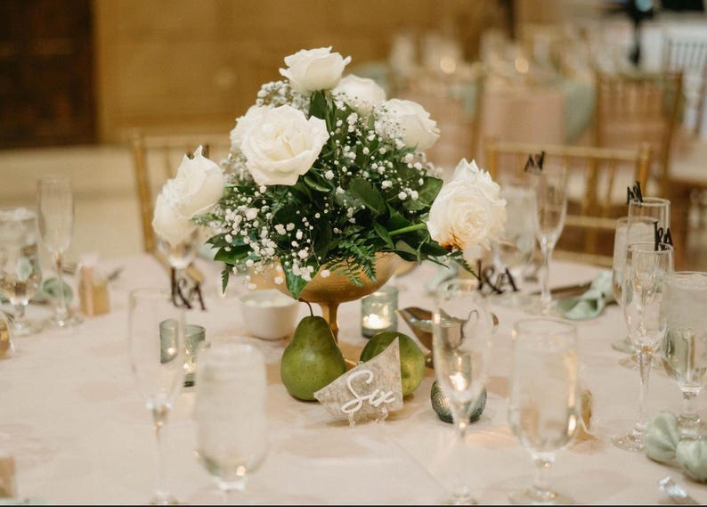 May include: A white floral centerpiece with white roses and baby's breath in a gold bowl. The centerpiece is on a white tablecloth with green placemats and clear glass glasses. The table number is "Six".
