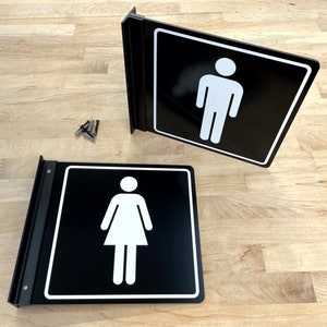Op de afbeelding: Twee zwart-witte toiletborden met een wit stickfiguur op een zwarte achtergrond. De borden zijn gemonteerd op een zwart metalen frame.