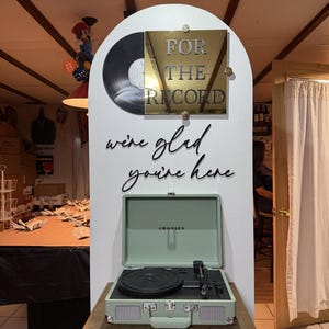 K&ouml;nnte beinhalten: Eine wei&szlig;e, bogenf&ouml;rmige Anzeigetafel zeigt eine schwarze Schallplatte und eine goldene quadratische Plakette mit dem Text FOR THE RECORD. Darunter steht in schwarzer Schreibschrift we're glad you're here. Ein hellgr&uuml;ner Plattenspieler steht davor.