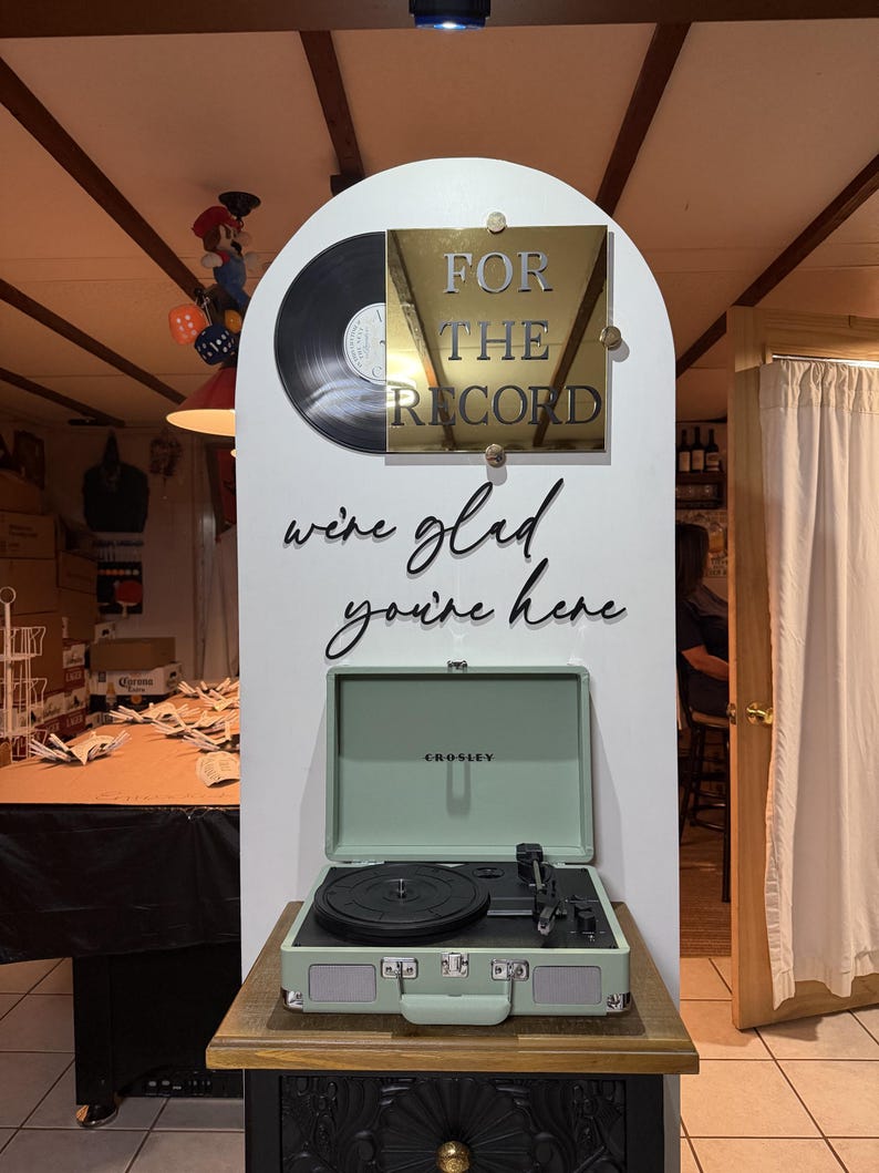 K&ouml;nnte beinhalten: Eine wei&szlig;e, bogenf&ouml;rmige Anzeigetafel zeigt eine schwarze Schallplatte und eine goldene quadratische Plakette mit dem Text FOR THE RECORD. Darunter steht in schwarzer Schreibschrift we're glad you're here. Ein hellgr&uuml;ner Plattenspieler steht davor.